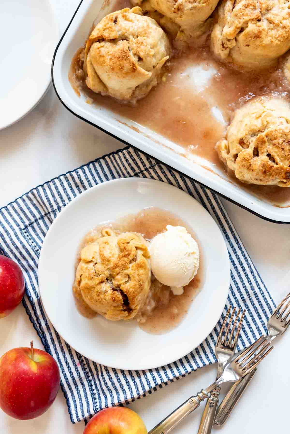 An apple dumpling on a plate with ice cream next to a pan with more apple dumplings.