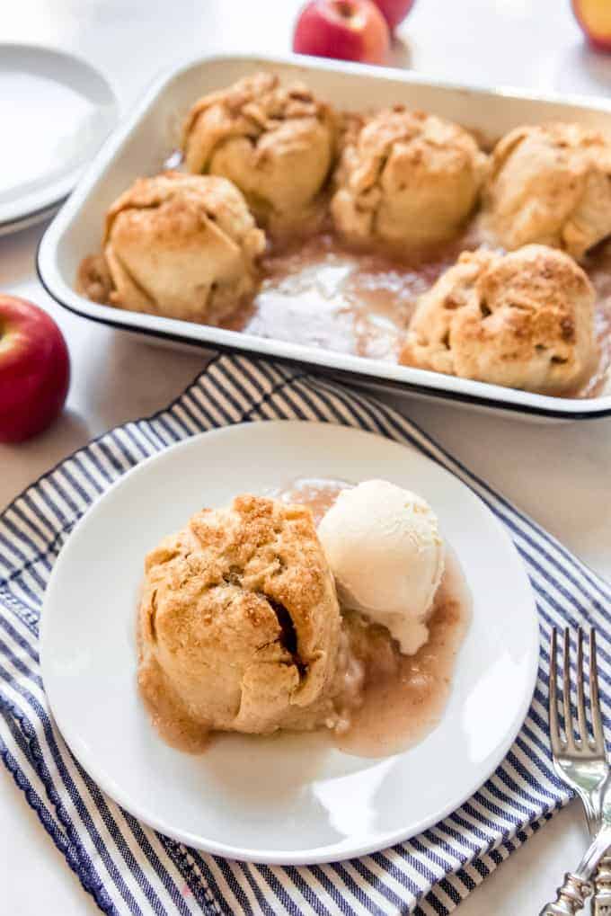 A baked apple dumpling on a plate next to a pan of more apple dumplings with red apples around them.