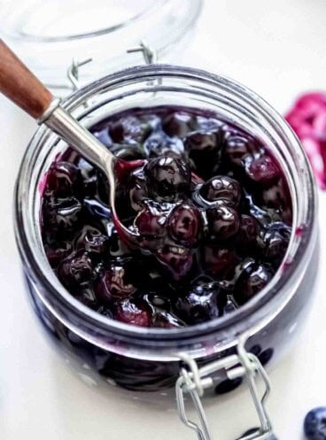 A glass jar of blueberry pie filling with a spoon lifting up some of the filling.