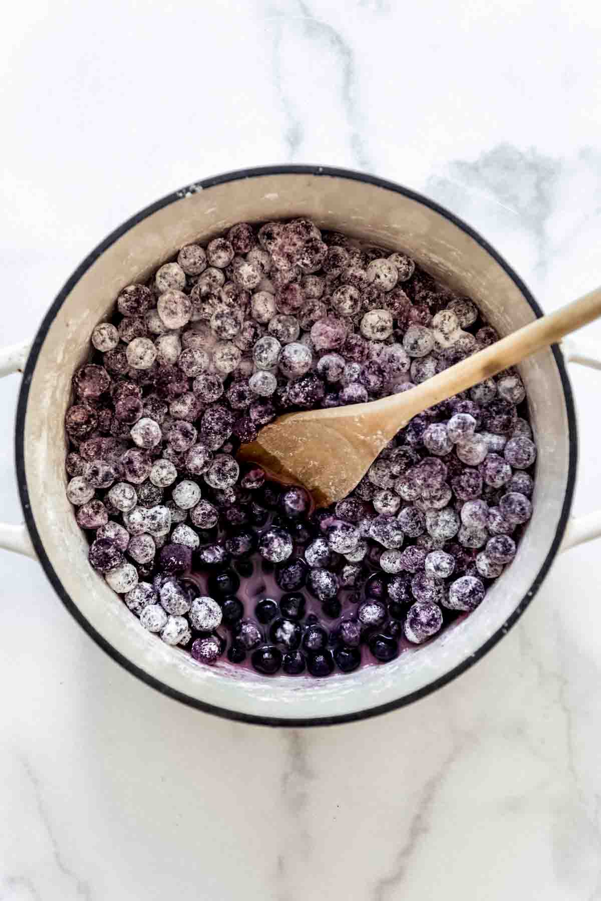 A large pot full of blueberries with sugar and cornstarch with a wooden spoon in it.