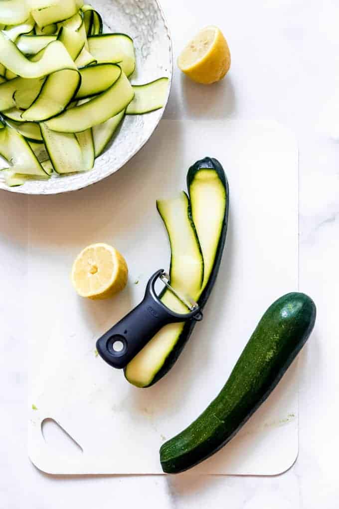 A zucchini being sliced into ribbons with a vegetable peeler on a white cutting board.