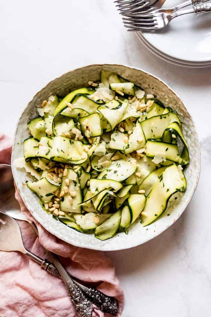 A large serving bowl of raw zucchini ribbons with pine nuts and parmesan next to a pink linen napkin and serving utensils.
