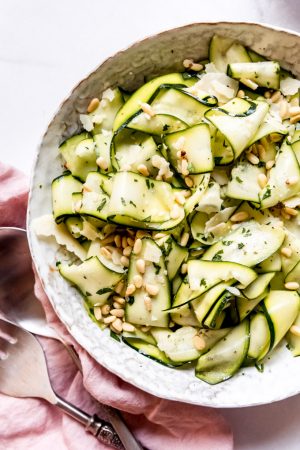 A large salad bowl filled with ribbons of raw zucchini salad sprinkled with pine nuts and parmesan shards.