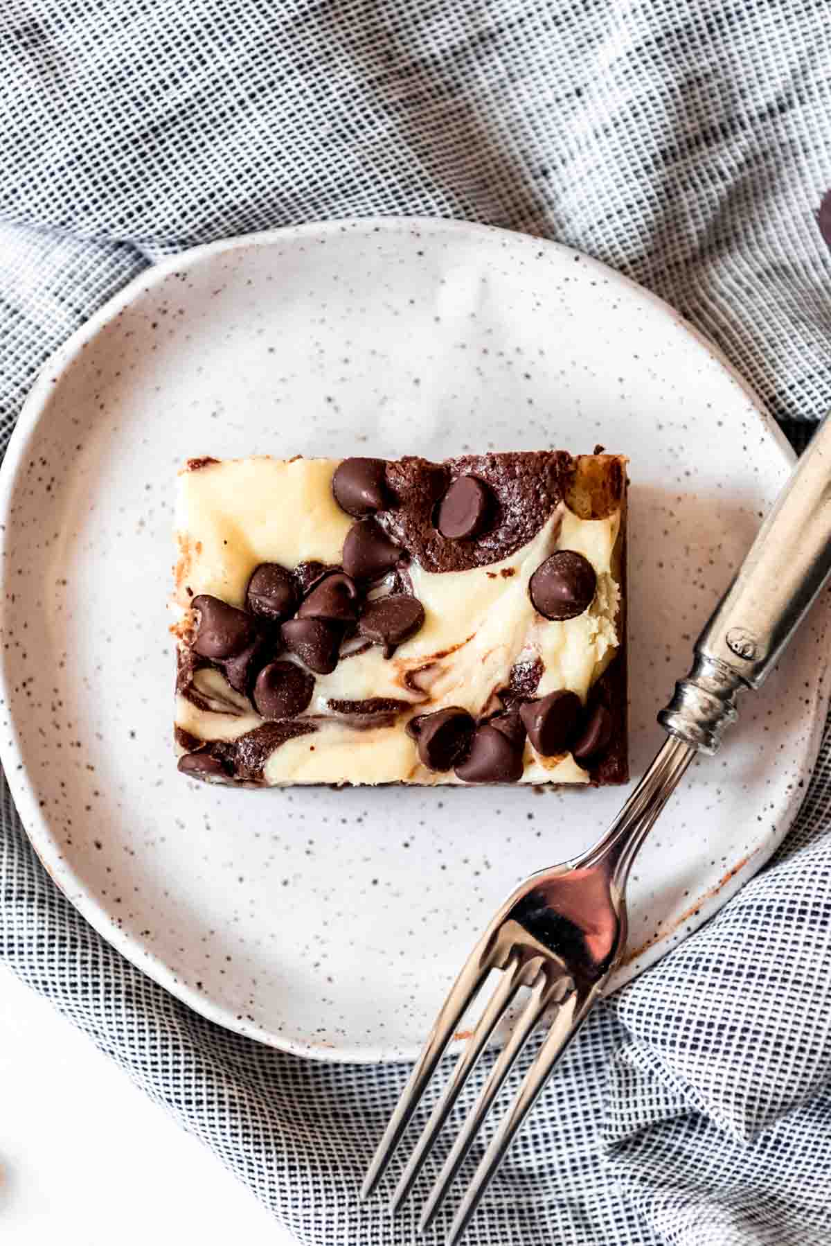 A slice of cream cheese brownie on a plate with a fork.