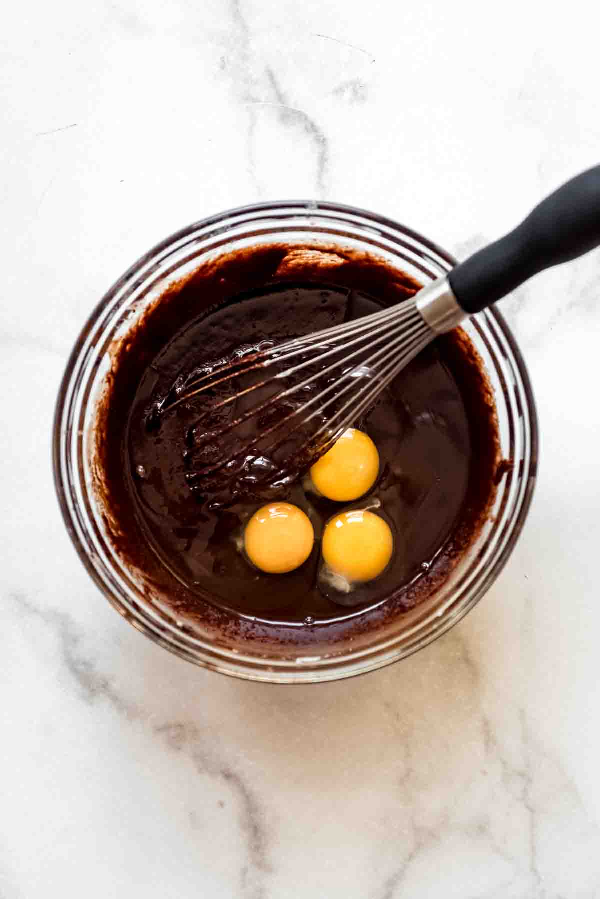 Adding 3 eggs to the brownie batter with a whisk.