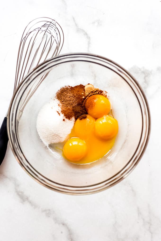 A glass bowl with sugar, pumpkin spices, and egg yolks next to a whisk.