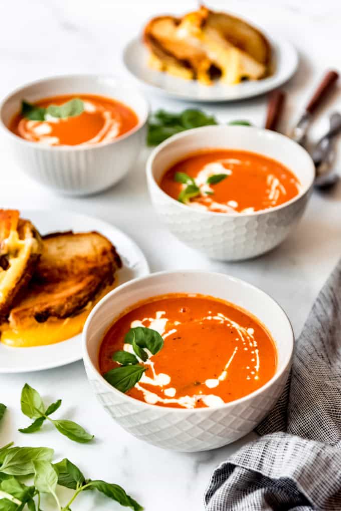 side shot of 3 tomato soup bowls drizzled with cream, with fresh basil and grilled cheese