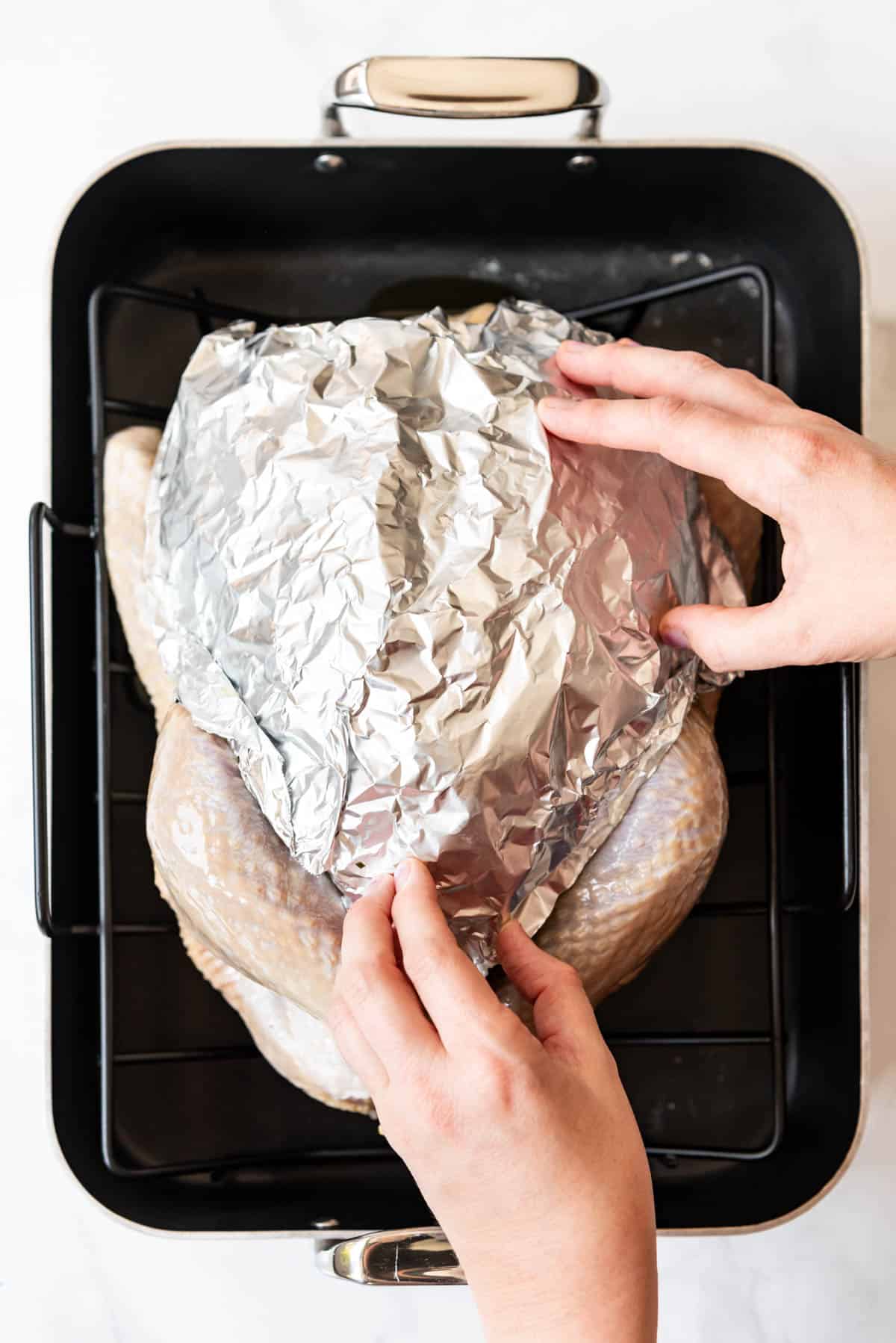 Hands molding a piece of aluminum foil around a turkey in a roasting pan.