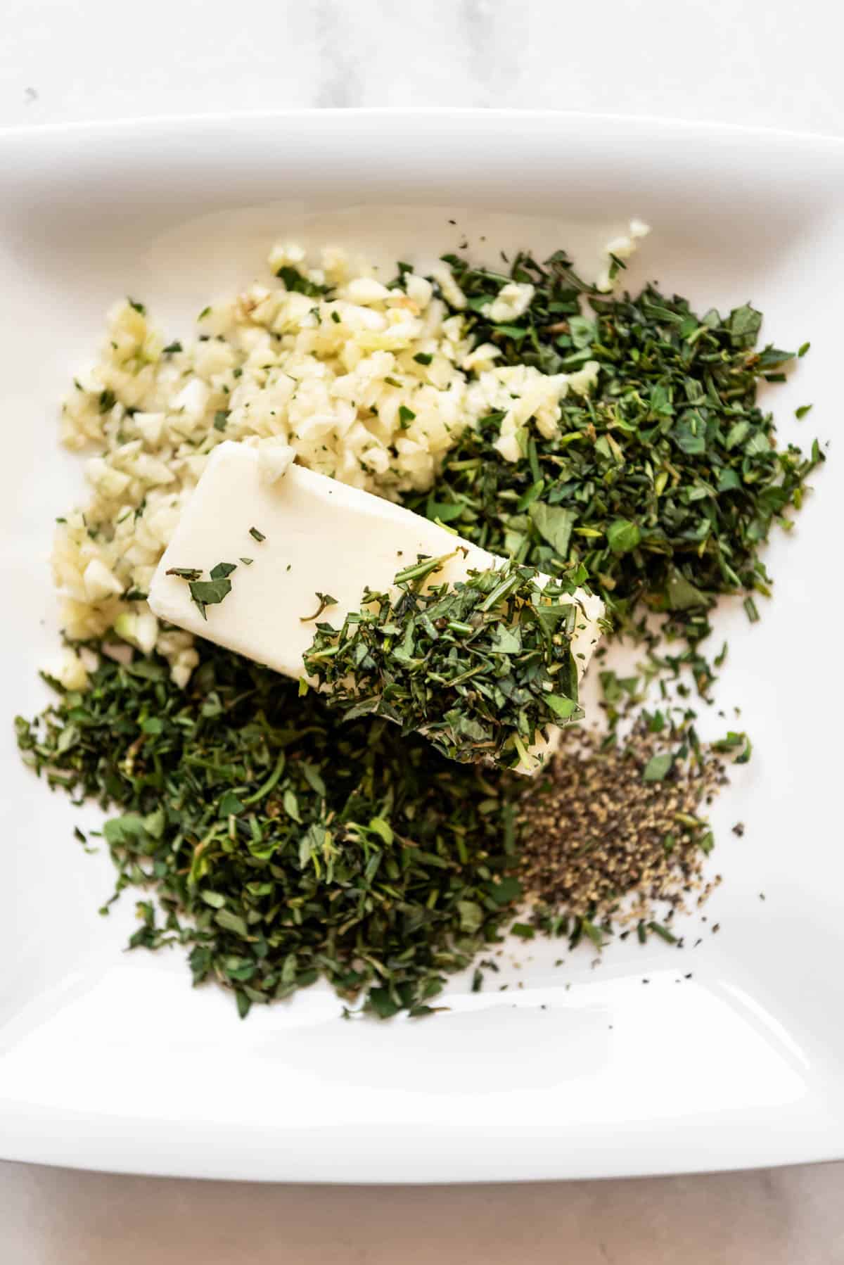 A stick of softened butter with chopped herbs and garlic on a plate.