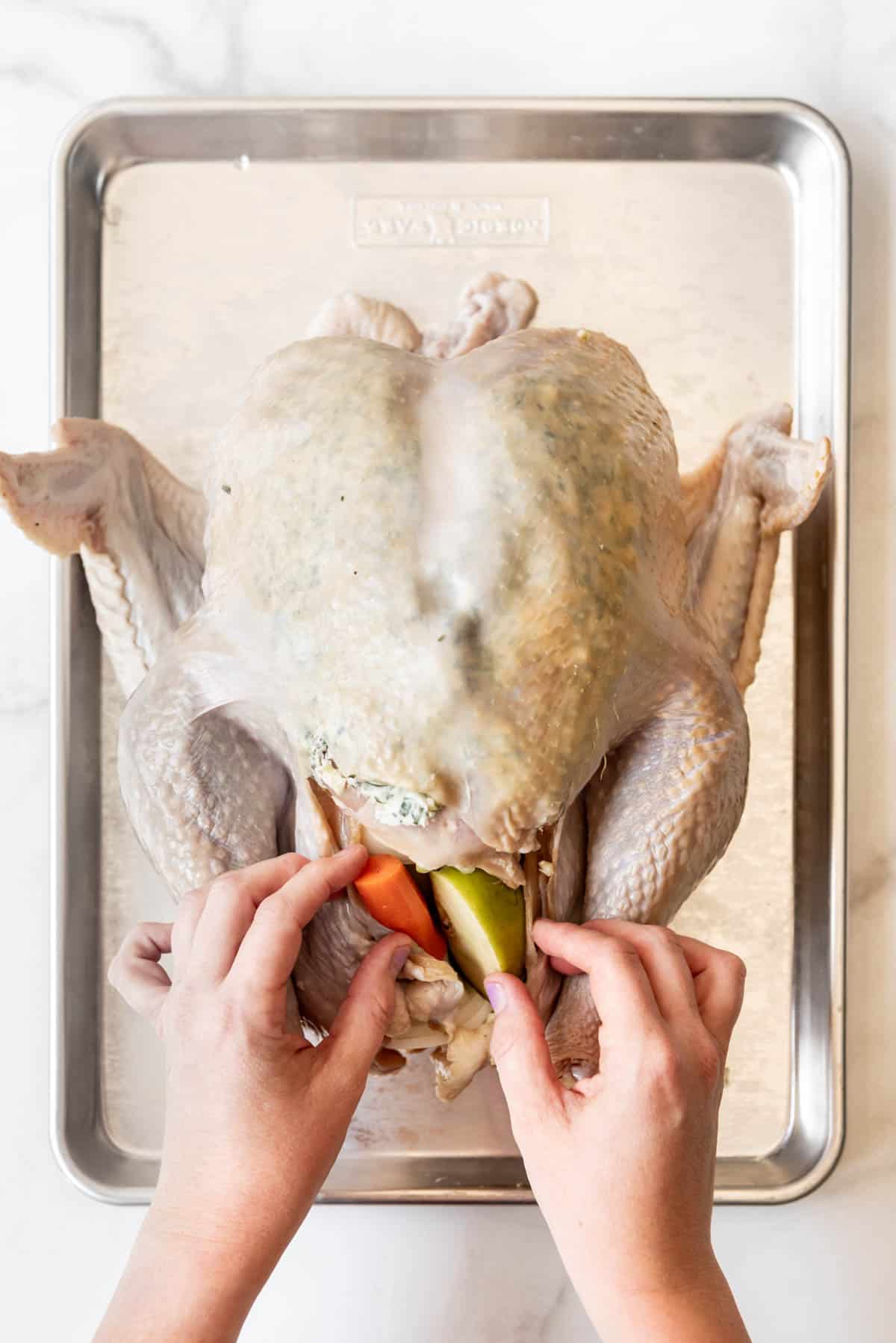 Hands stuffing aromatics into the cavity of a large turkey on a baking sheet.