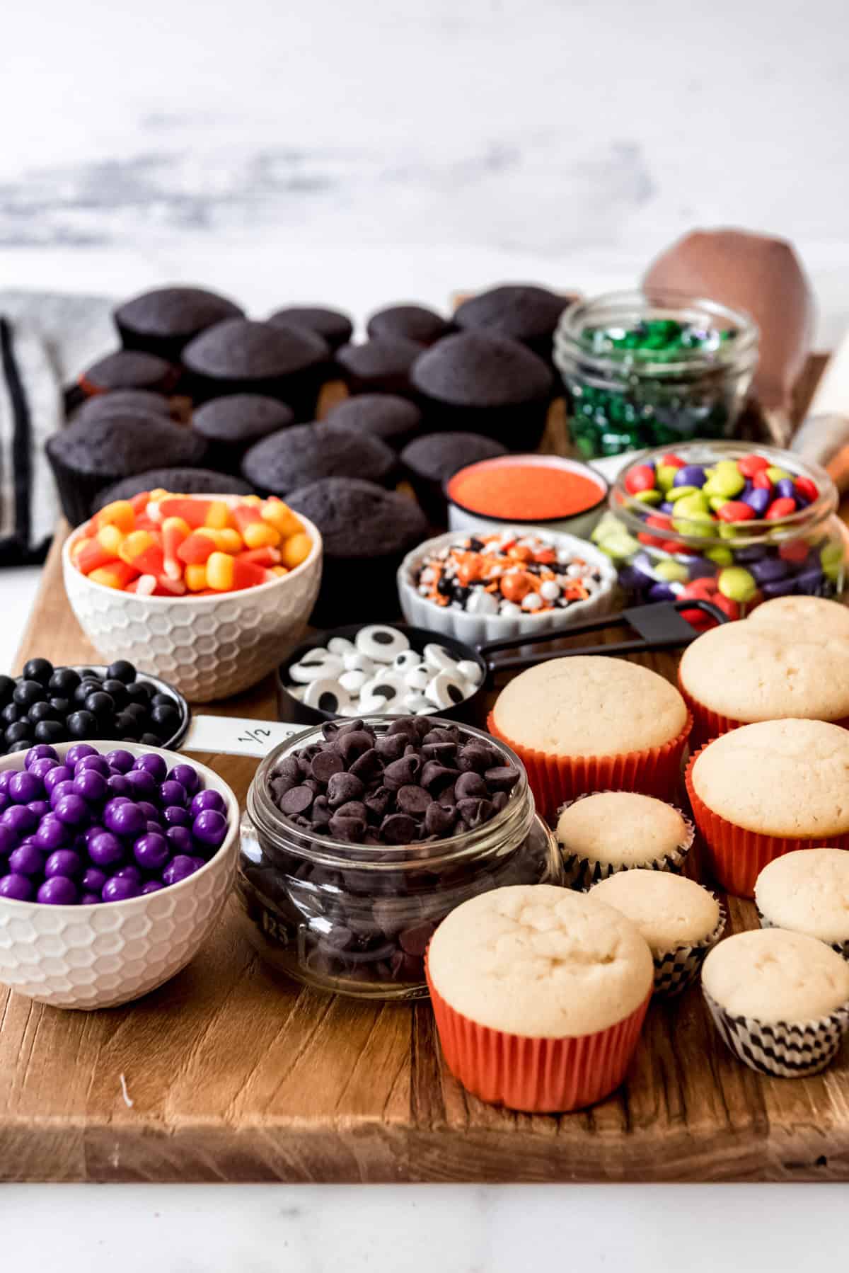 Jars of chocolate chips, candy, and sprinkles on a board next to unfrosted cupcakes.