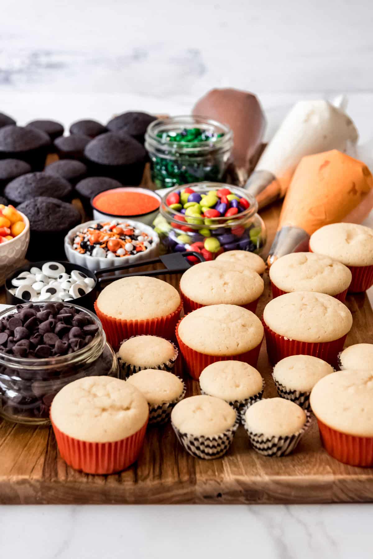 Vanilla cupcakes in orange, black, and white wrappers on a board with candy and frosting for decorating.