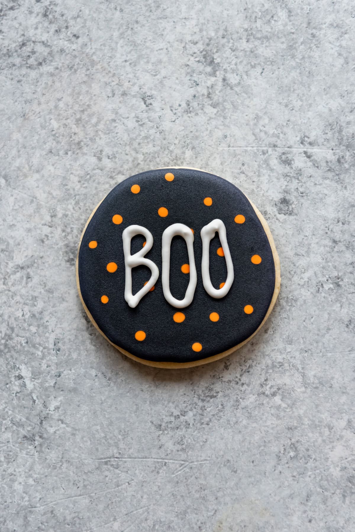 A decorated Halloween sugar cookie with orange dots on a black background and the word "boo" in white icing.