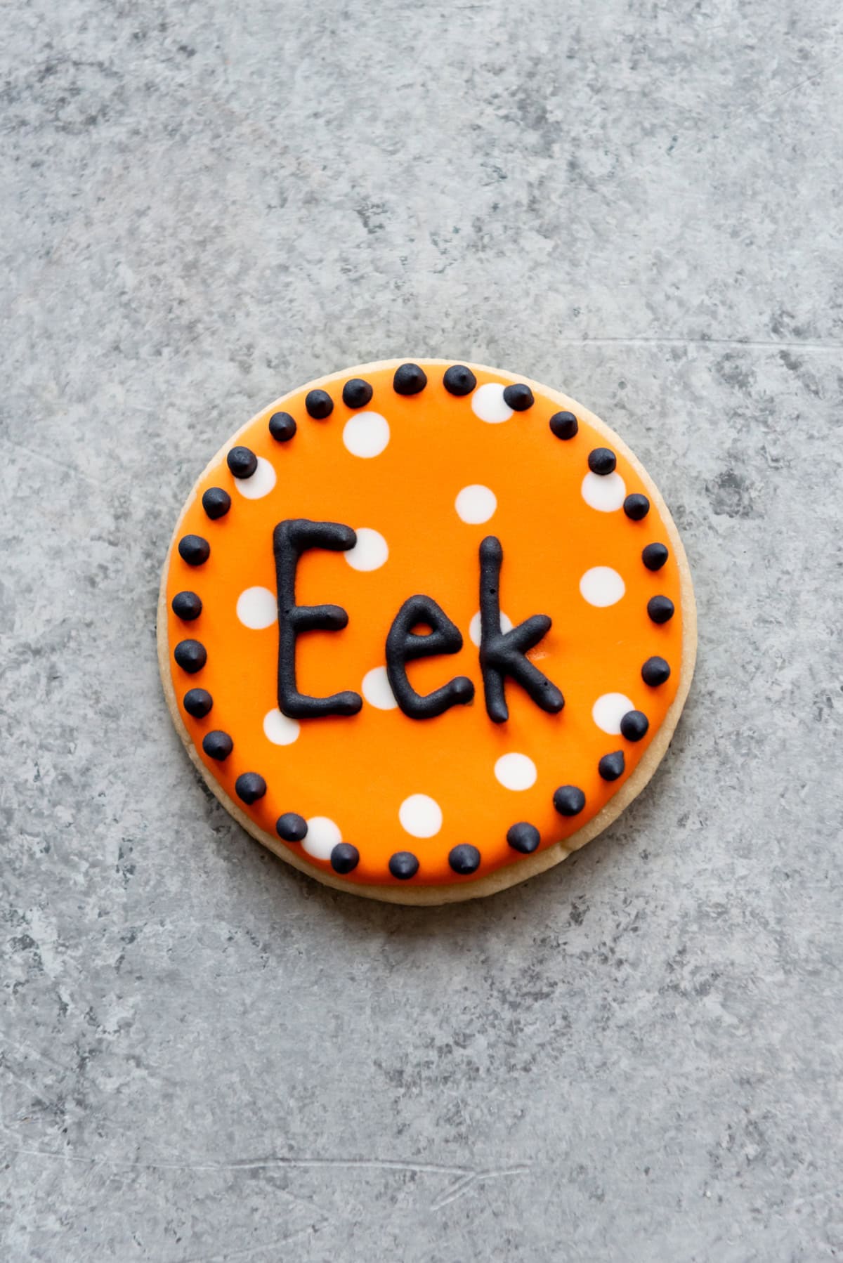 A sugar cookie with white dots on an orange background with the word "eek" piped in black icing.