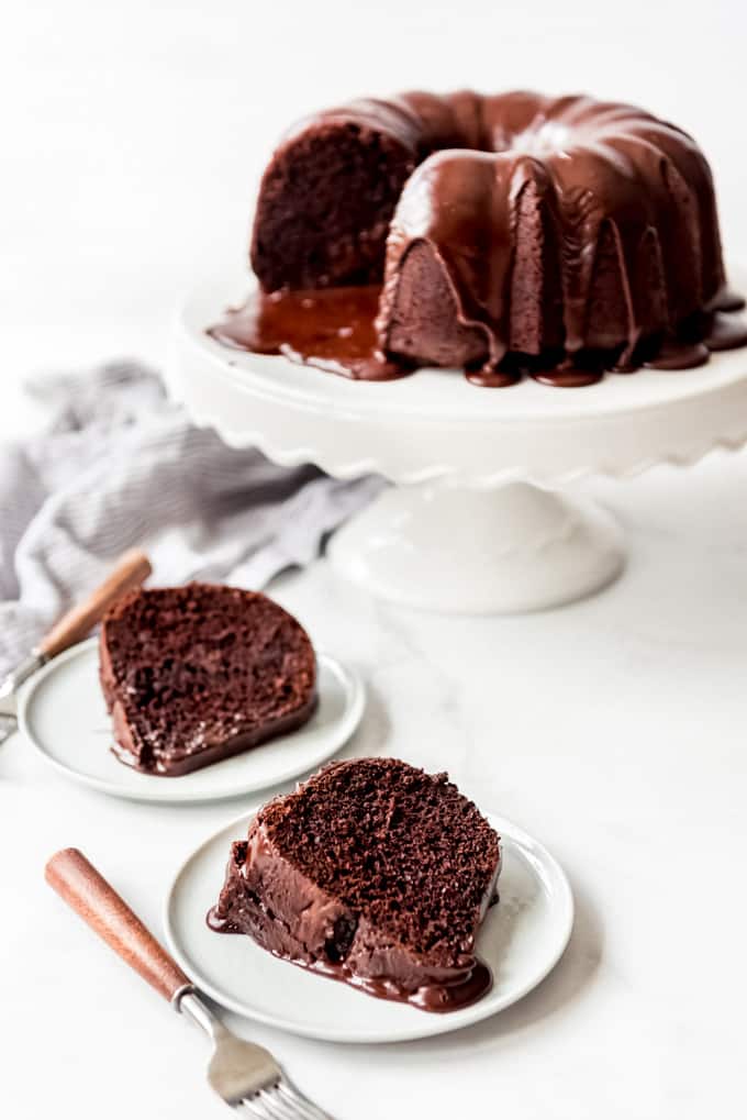 2 slices of chocolate bundt cake on a whites plate with rest of cake in background on cakestand