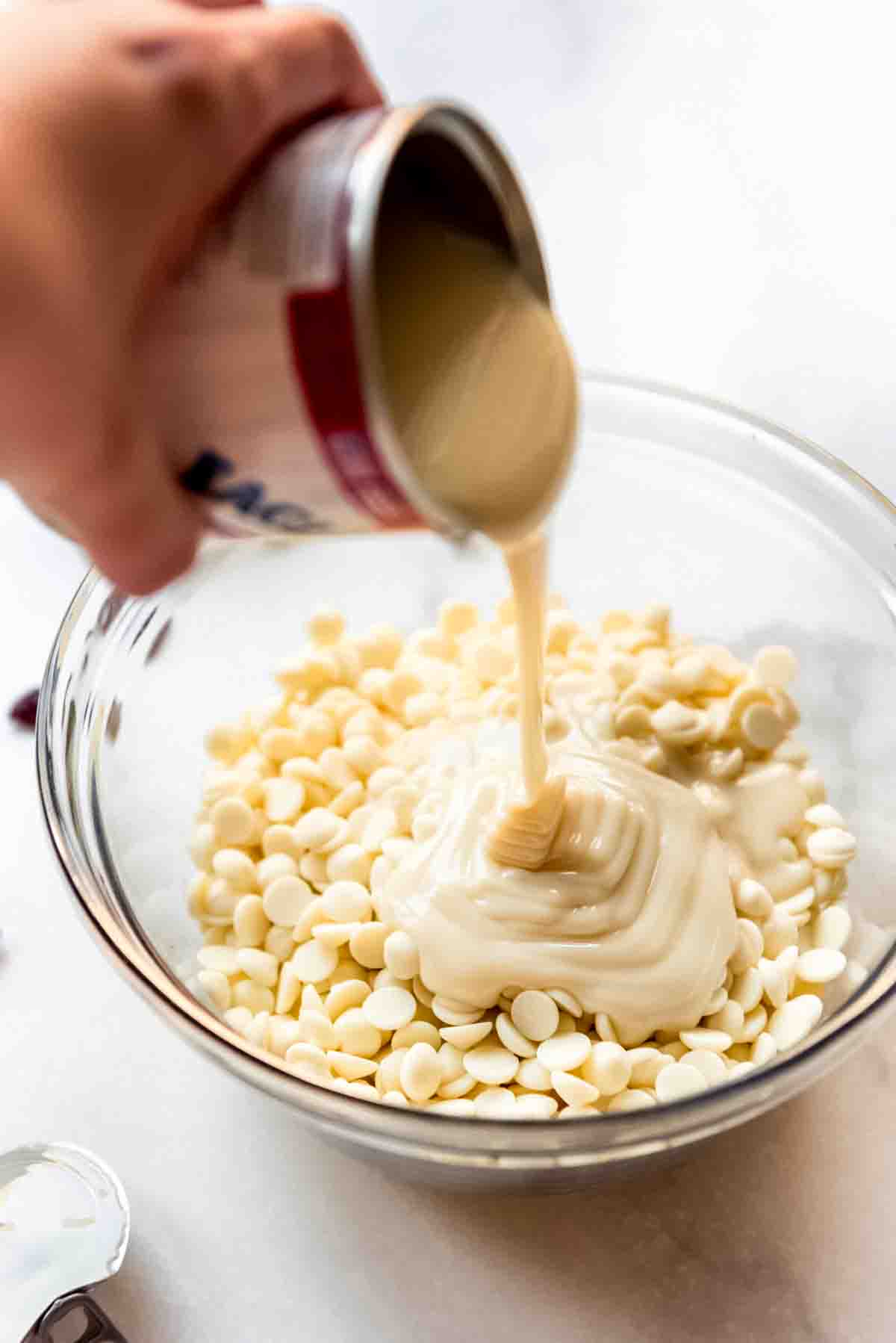 Sweetened condensed milk is poured on top of white chocolate chips in a clear glass bowl. 