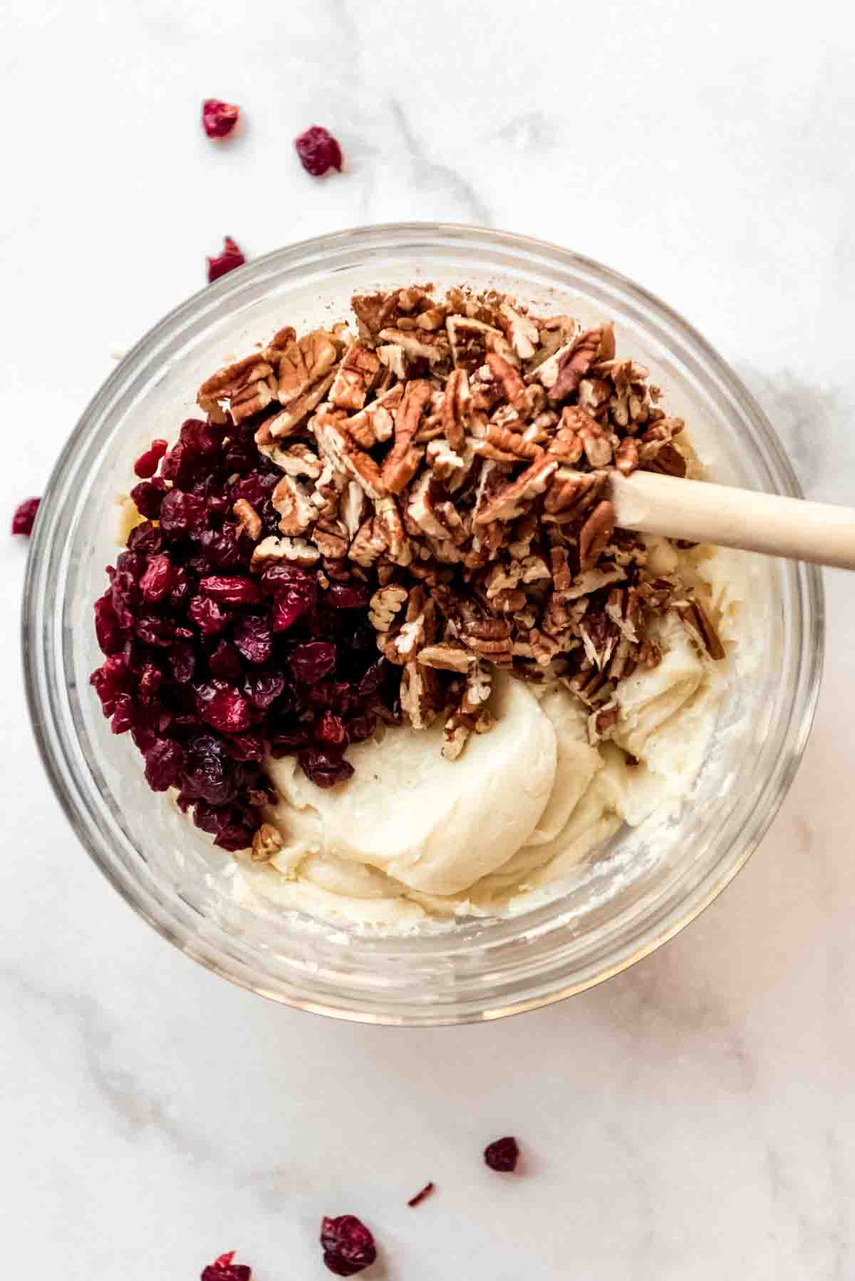 Dried cranberries and chopped pecans are added on top of the white chocolate mixture in a clear glass bowl. 