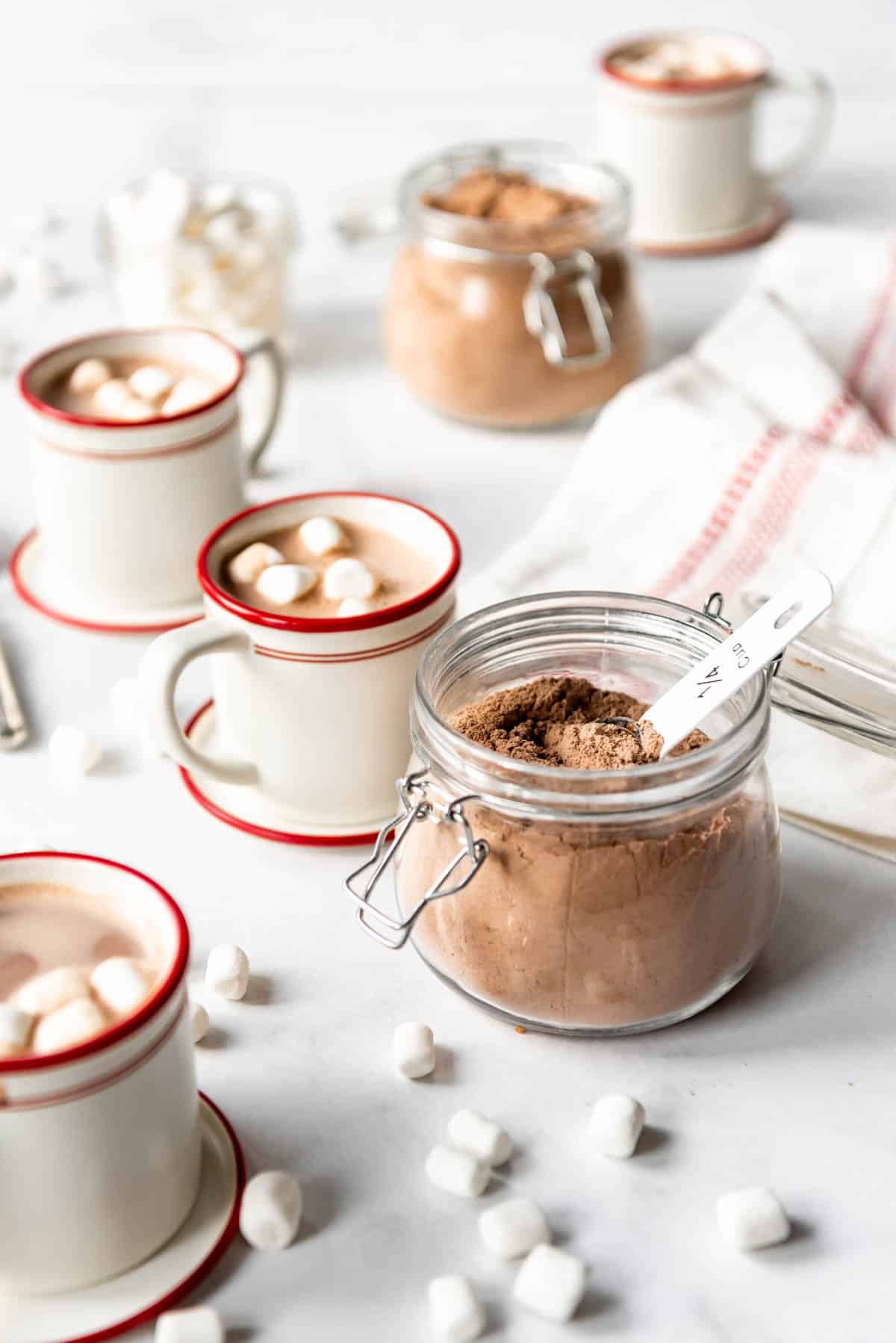 A jar of hot chocolate mix next to mugs of hot chocolate.