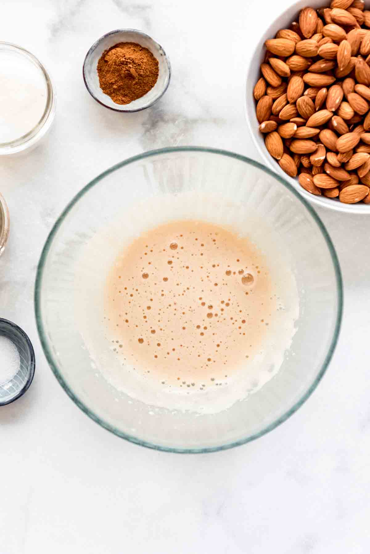 Vanilla and egg white beaten together until foamy, in a glass bowl. Other bowls are visible around this, containing almonds and other ingredients. 