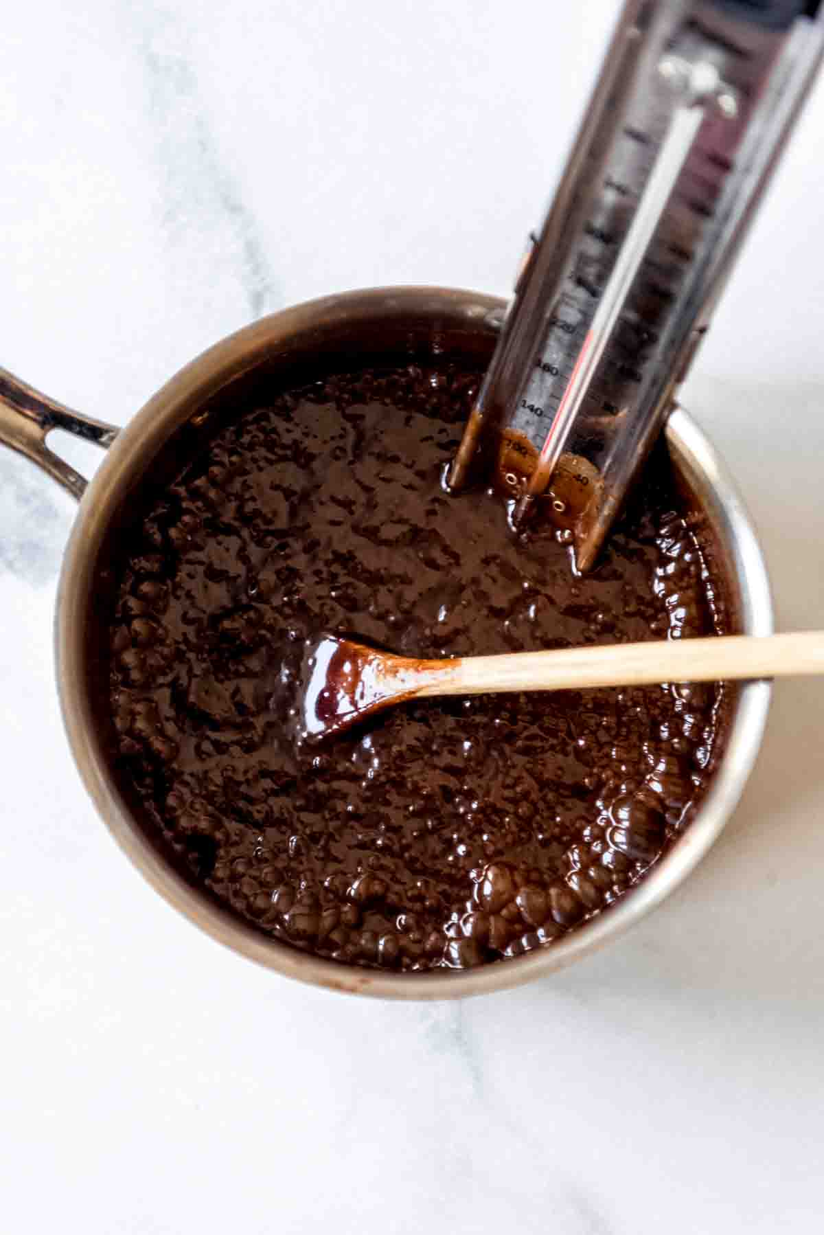 Praline mixture is boiling in metal pot, with candy thermometer and wooden spoon placed inside the mixture. 