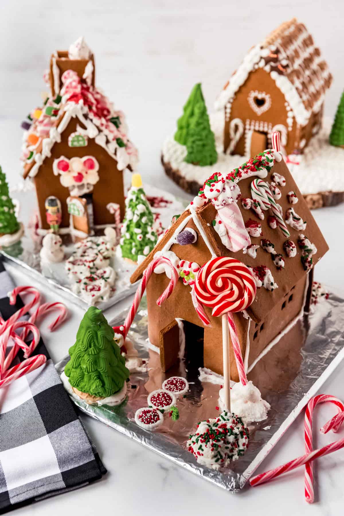 Three completed gingerbread houses are decorated with candy and rest on a white surface. Candy canes and a checkered cloth are visible around the decorated gingerbread houses. 