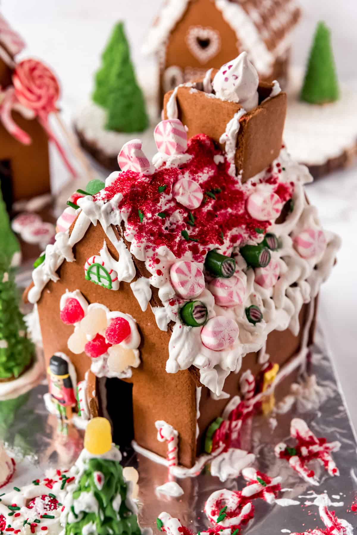 An overhead view of the decorated roof of a holiday gingerbread house with a chimney and candy. Two decorated gingerbread houses are visible in the background. 