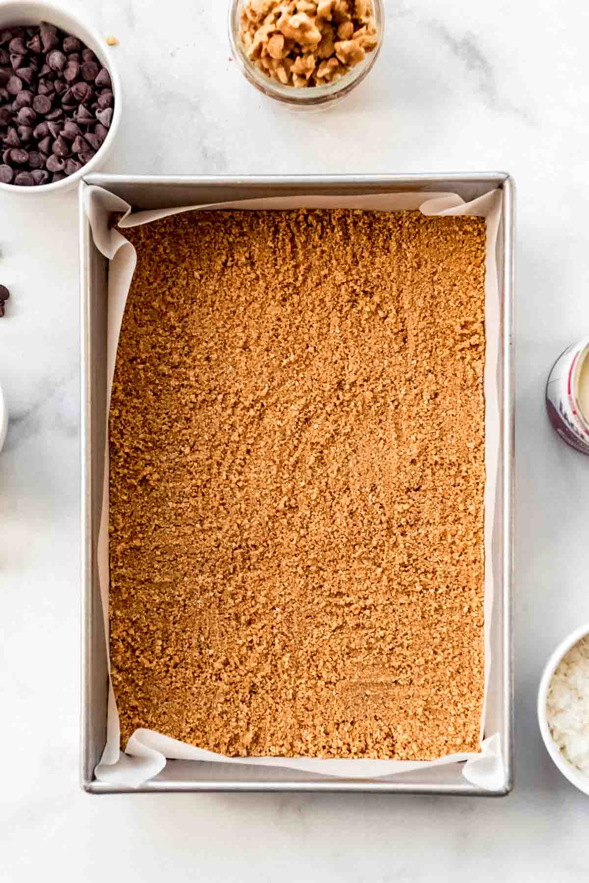 A crushed graham cracker and melted butter crust has been evenly pressed into a rectangle pan prepared with a parchment paper sling. 