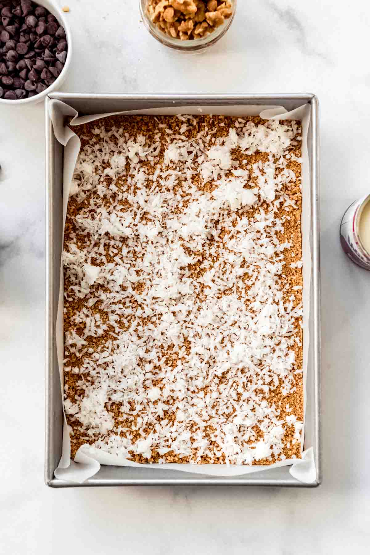 Coconut flakes are sprinkled on top of a graham cracker crust in a 9 by 13 inch baking dish. Bowls of chocolate chips and nuts are visible around the dish. 