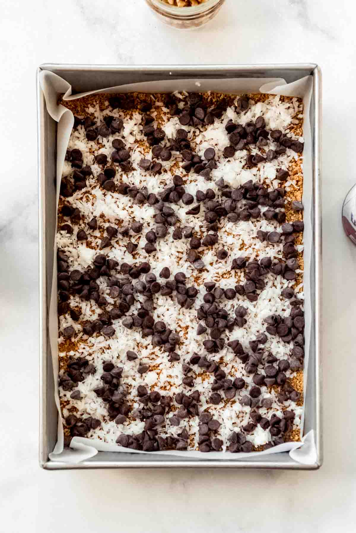 Chocolate chips are scattered on top of coconut and graham cracker layers in a baking dish. 