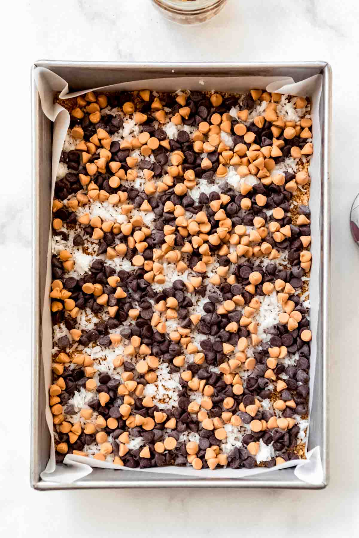 Butterscotch chips are added to a pan filled with chocolate chips, shredded coconut and a graham cracker crust. 