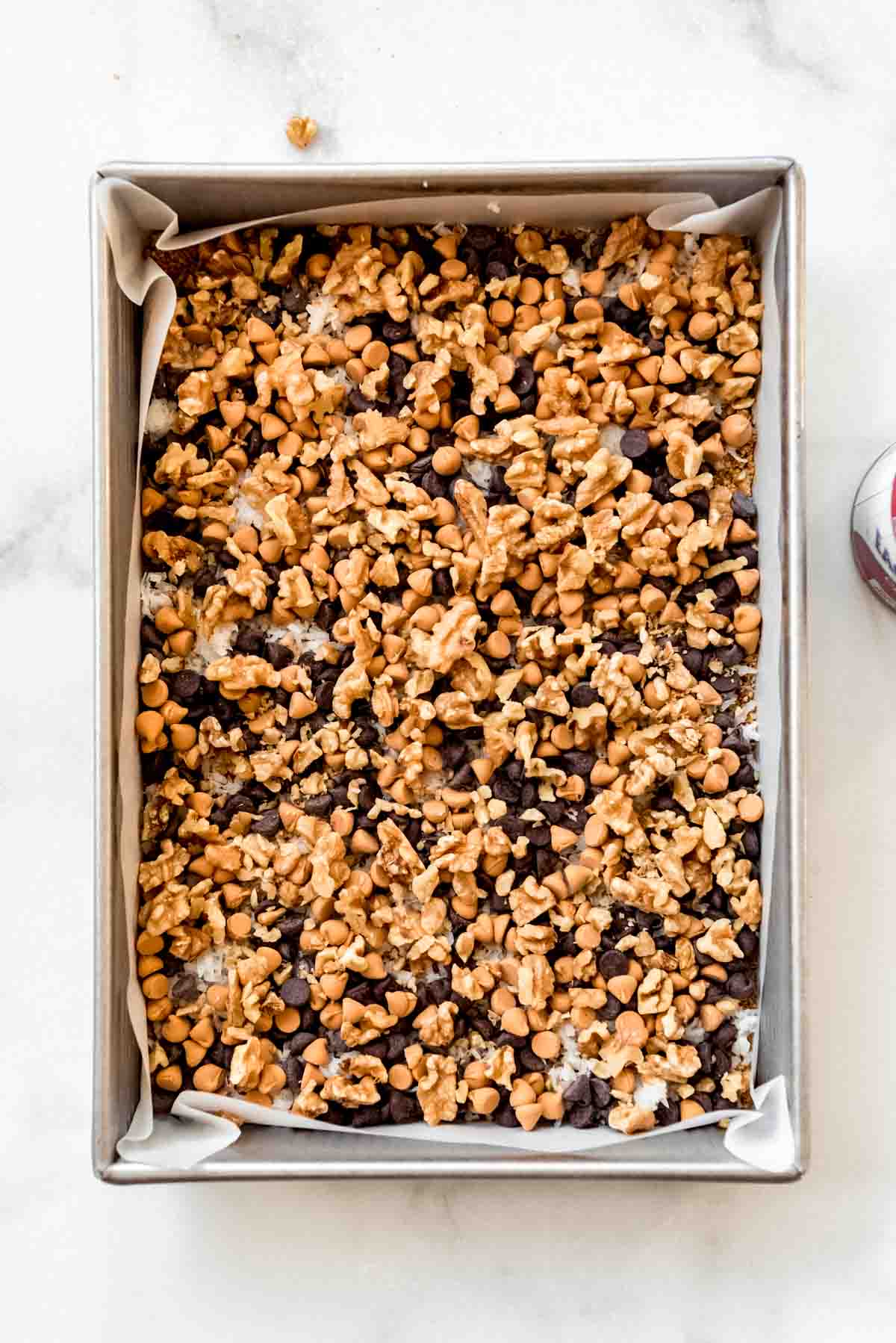 Chopped walnuts are scattered on top of butterscotch and chocolate chips, with some coconut flakes visible, all contained in a metal 9 by 13 inch baking dish. 