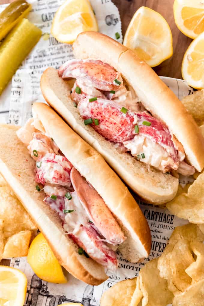 Two lobster rolls next to chips and lemons.