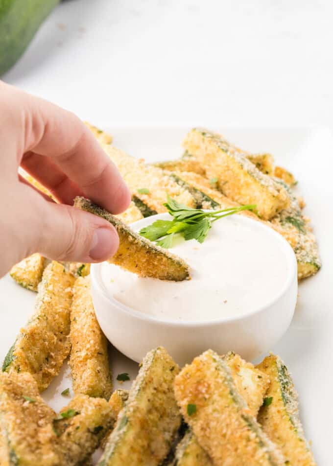A hand dipping zucchini fries into a bowl.