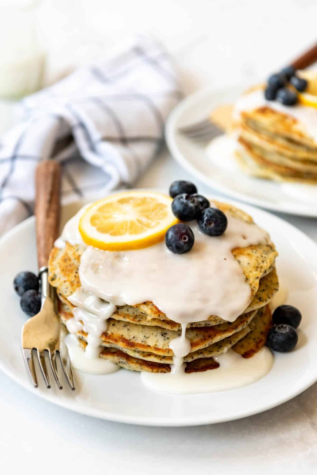 A stack of lemon poppy seed pancakes on a plate with a lemon slice and blueberries on top.