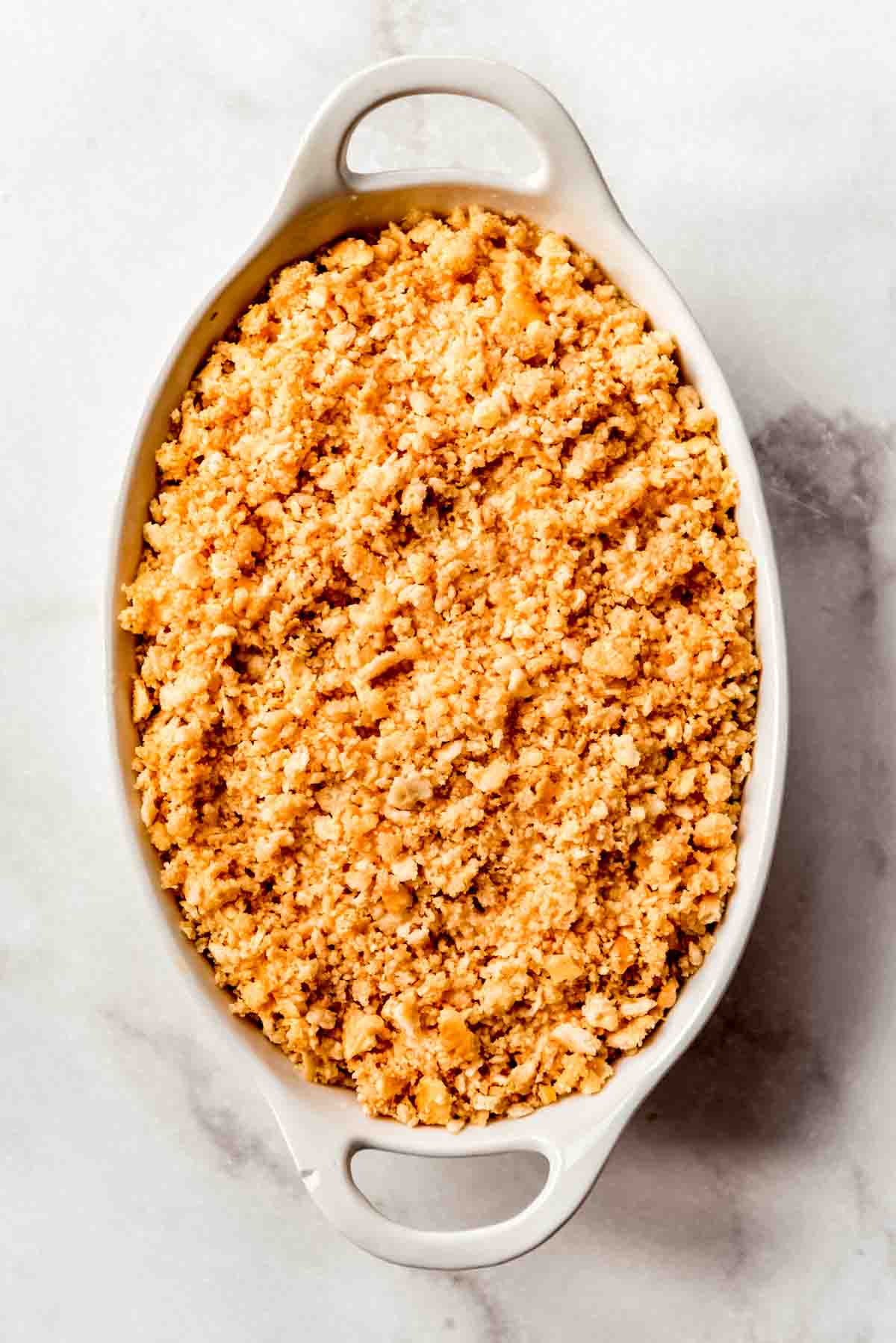 A cracker crumb topping is placed on top of a pineapple casserole in a white oval casserole dish. 