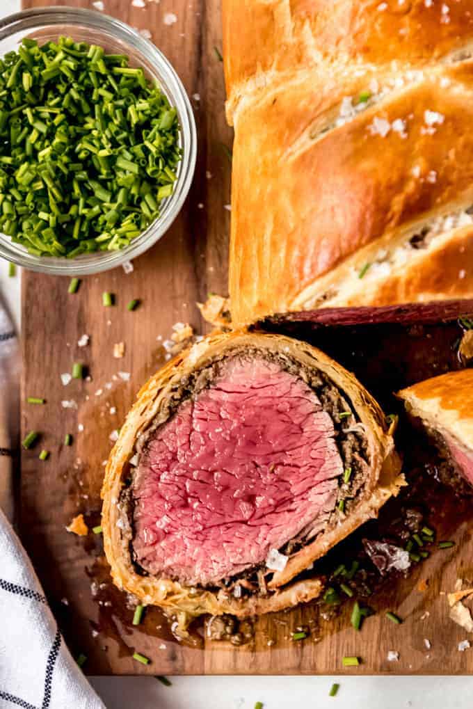 medium rare beef wellington slice on wooden board with chives and rest of beef wellington.