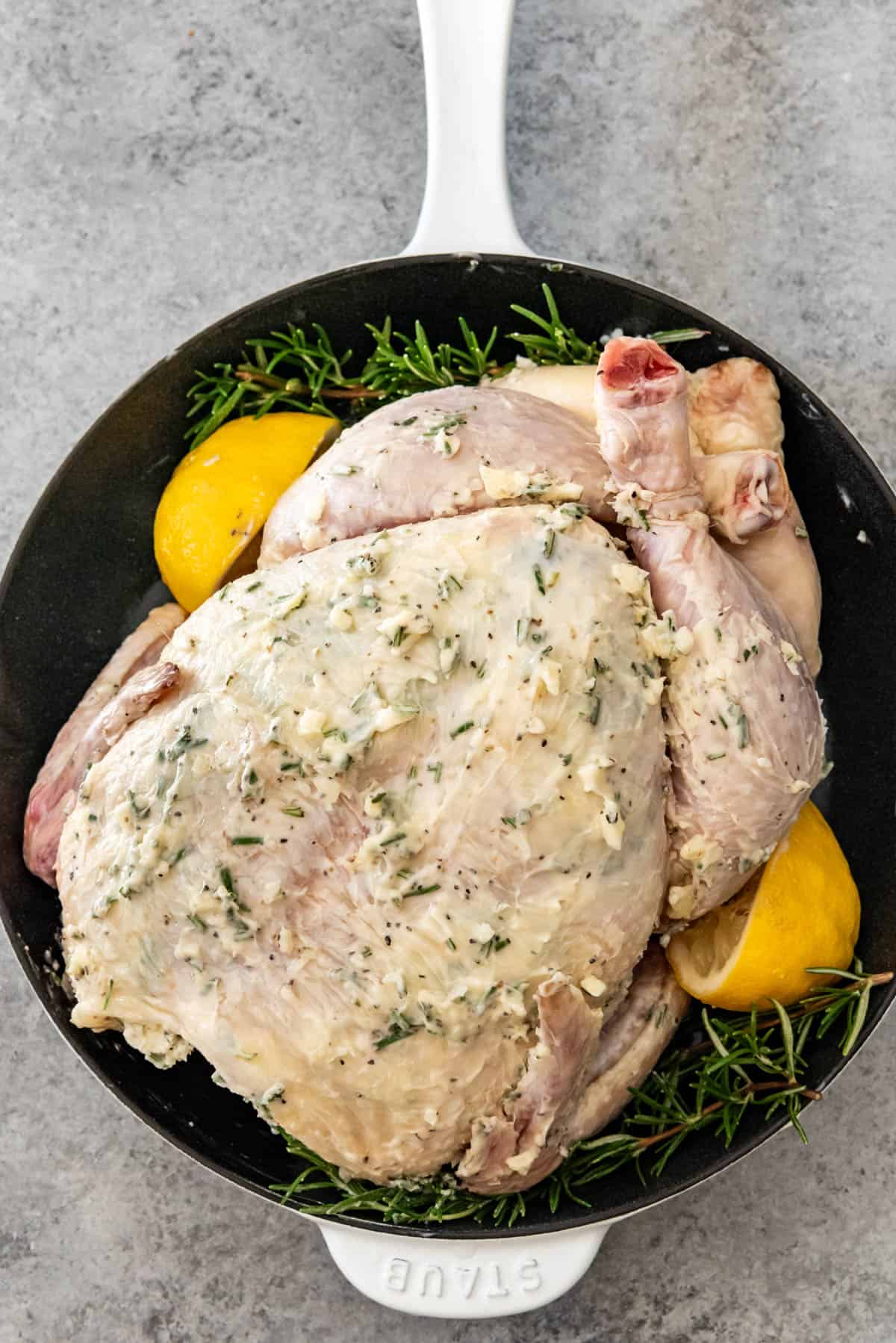 A whole chicken in roasting pan (dutch oven) covered in garlic butter, beside rosemary and lemon.