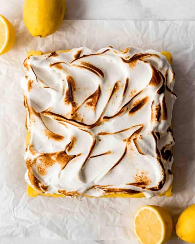 Toasted meringue is visible on top of the lemon pie bar base, resting on white parchment paper. Whole and cut lemons are scattered around the dessert.