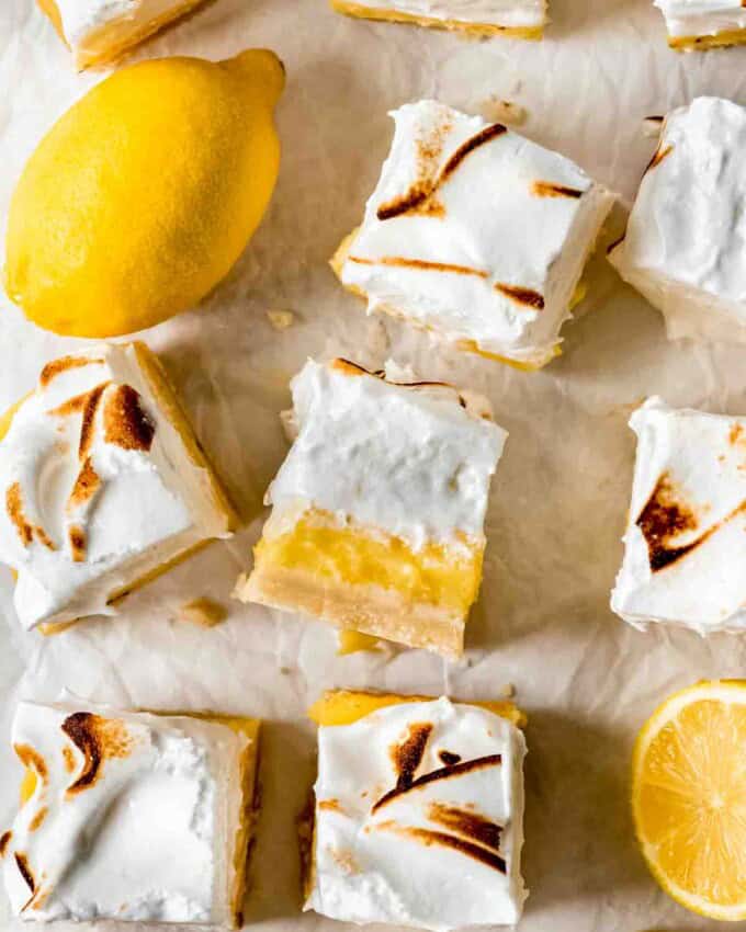 Squares of cut lemon meringue pie bars are scattered on white parchment paper. One is turned on its side to show the crust, filling and topping of the bars, and cut and whole lemons are sitting among the bars.