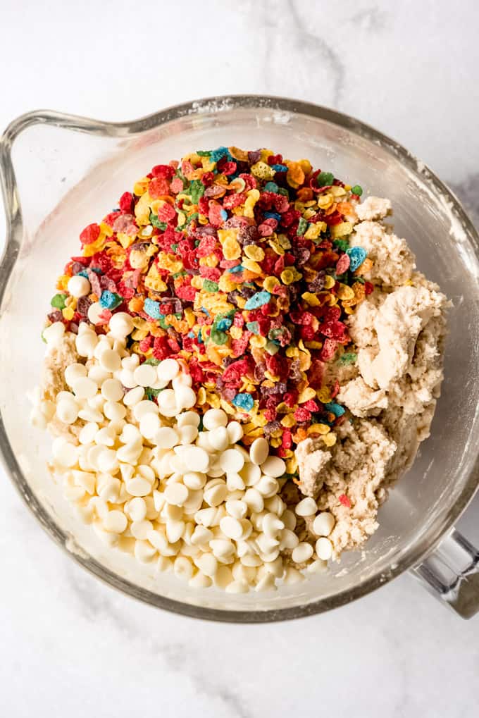 Adding Fruity Pebbles and white chocolate chips to cookie dough in a bowl.