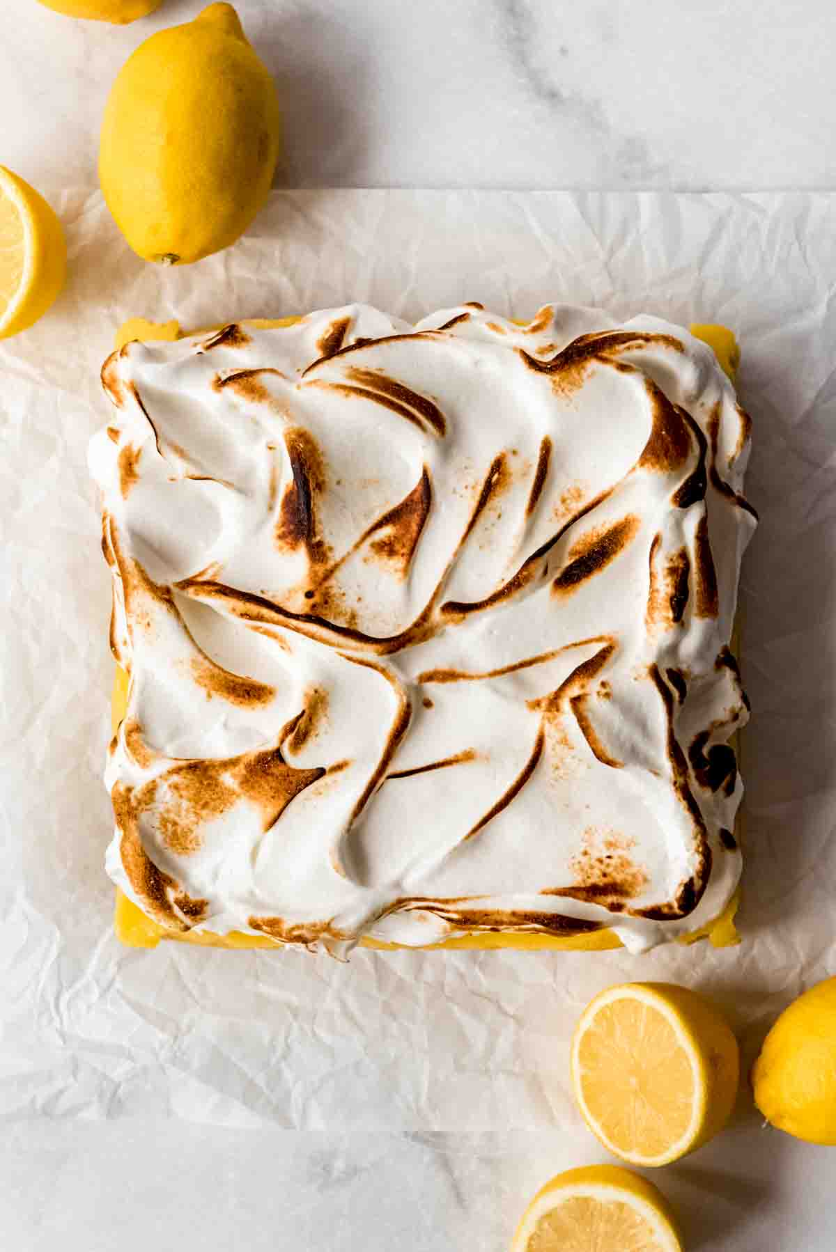 Toasted meringue is visible on top of the lemon pie bar base, resting on white parchment paper. Whole and cut lemons are scattered around the dessert. 