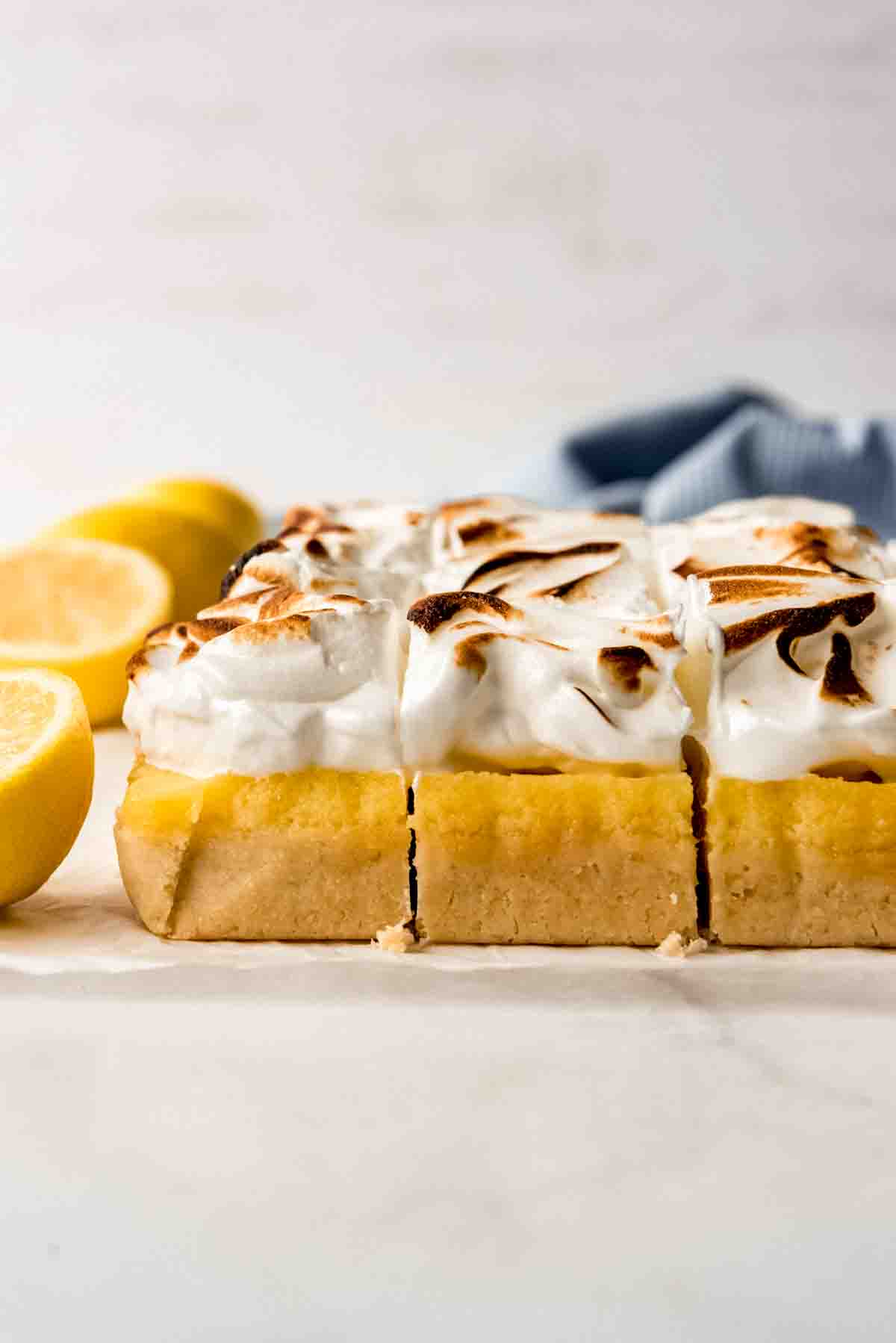 Cut lemon meringue pie bars are lined up neatly on white parchment paper. Cut lemons are visible in a line on the left side of the image, and a blue cloth is visible in the background. 