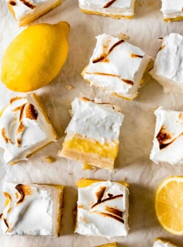 Squares of cut lemon meringue pie bars are scattered on white parchment paper. One is turned on its side to show the crust, filling and topping of the bars, and cut and whole lemons are sitting among the bars.