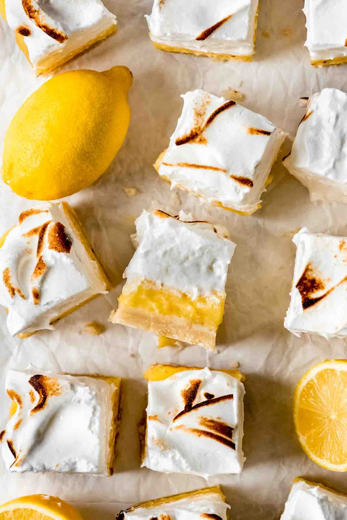 Squares of cut lemon meringue pie bars are scattered on white parchment paper. One is turned on its side to show the crust, filling and topping of the bars, and cut and whole lemons are sitting among the bars. 