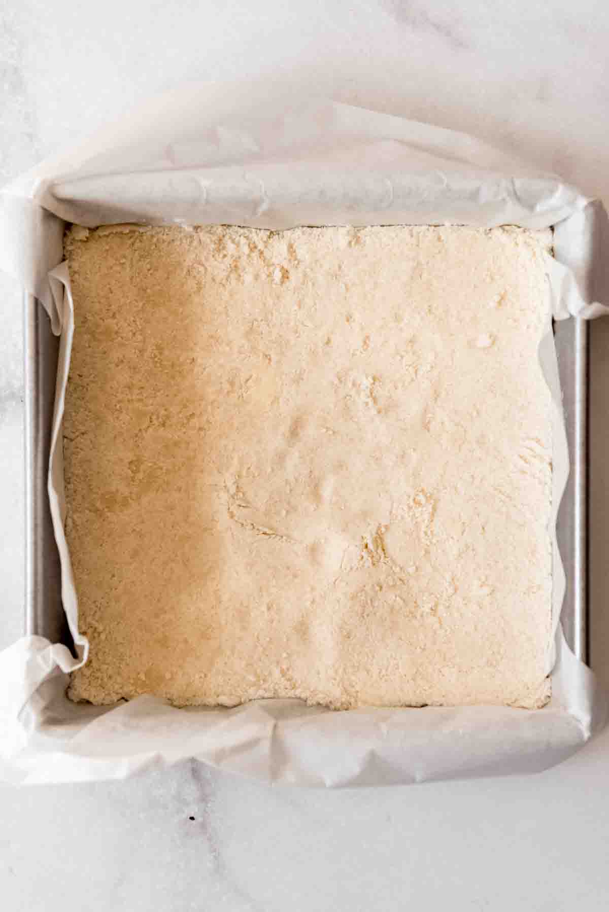 A crust mixture has been pressed into the bottom of a square baking pan with a parchment paper sling 