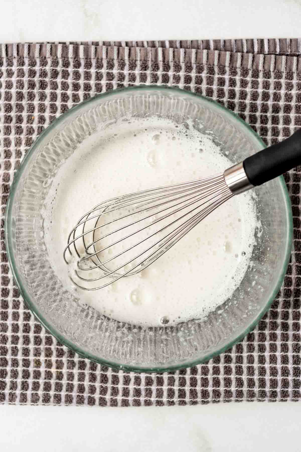 Ingredients for  meringue topping are being mixed with a whisk in a glass bowl. 