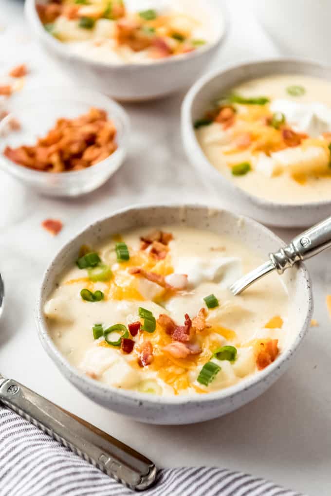 3 bowls of loaded baked potato soup with garnishes in background and silverware at side
