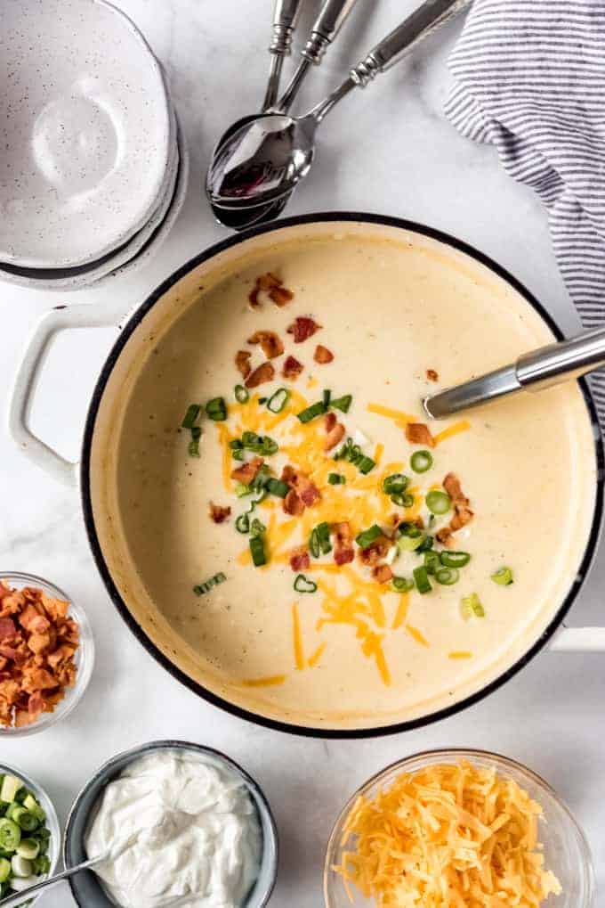 overhead, large pot of Loaded Baked Potato Soup with garnishes in separate small bowls, and pile of empty bowls next to silverware