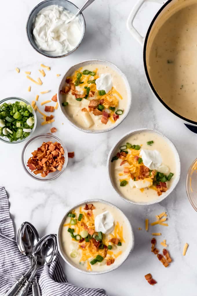 overhead shot of 3 bowls of loaded baked potato soup with garnishes in background and spoons at side, and large pot of soup