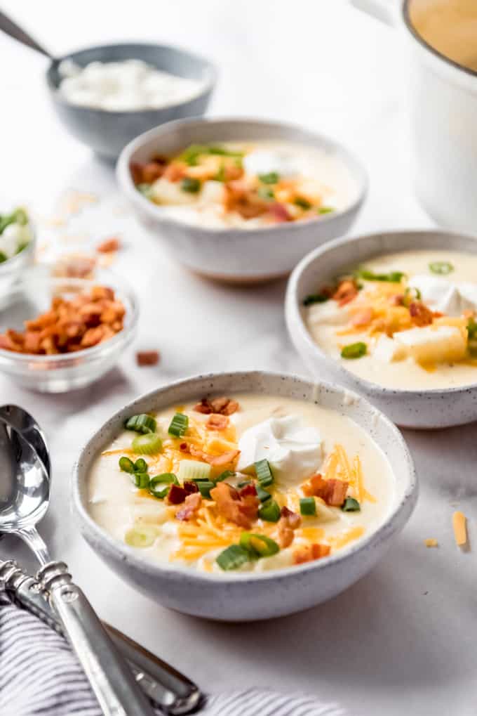 3 bowls of loaded baked potato soup with garnishes in background and spoons at side